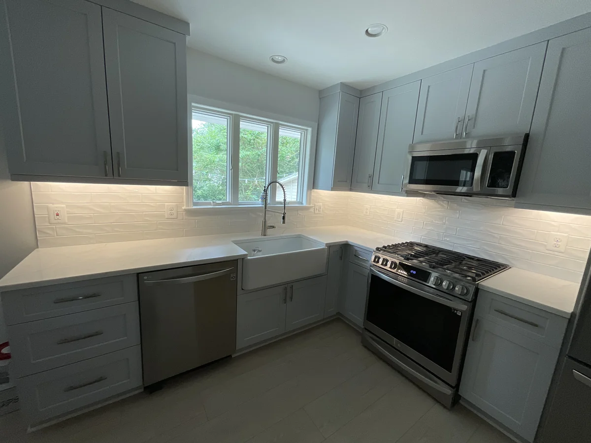 Light stone countertops and a white tile backsplash around gray kitchen cabinets and a farmhouse sink