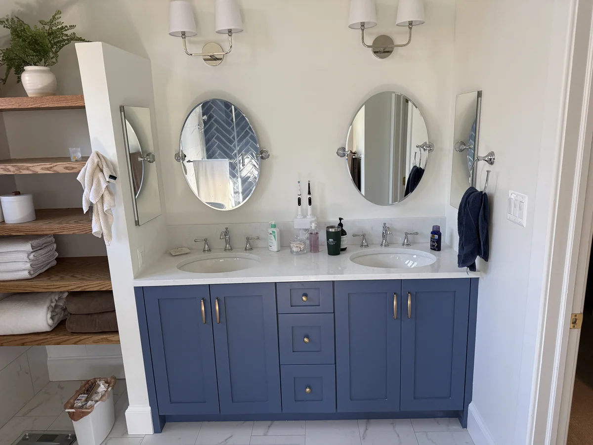 Blue double vanity with light stone countertops, oval mirrors and porcelain floor tile.